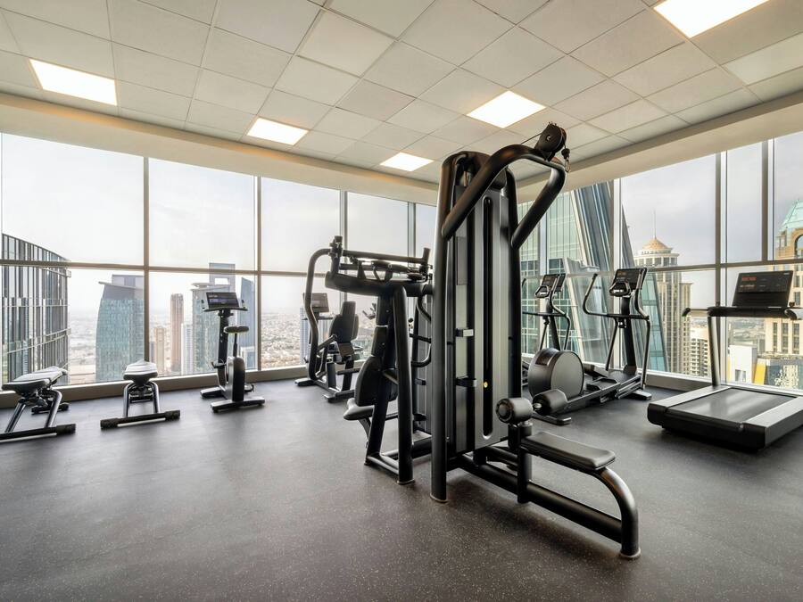Un gimnasio con diverso equipamiento de ejercicio, ventanas amplias y vistas a un paisaje urbano.