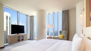 A hotel room with a large bed, a flat-screen TV on a wooden cabinet, a chair, and a view of the cityscape through large windows. - Le Méridien City Center, Doha (Doha)