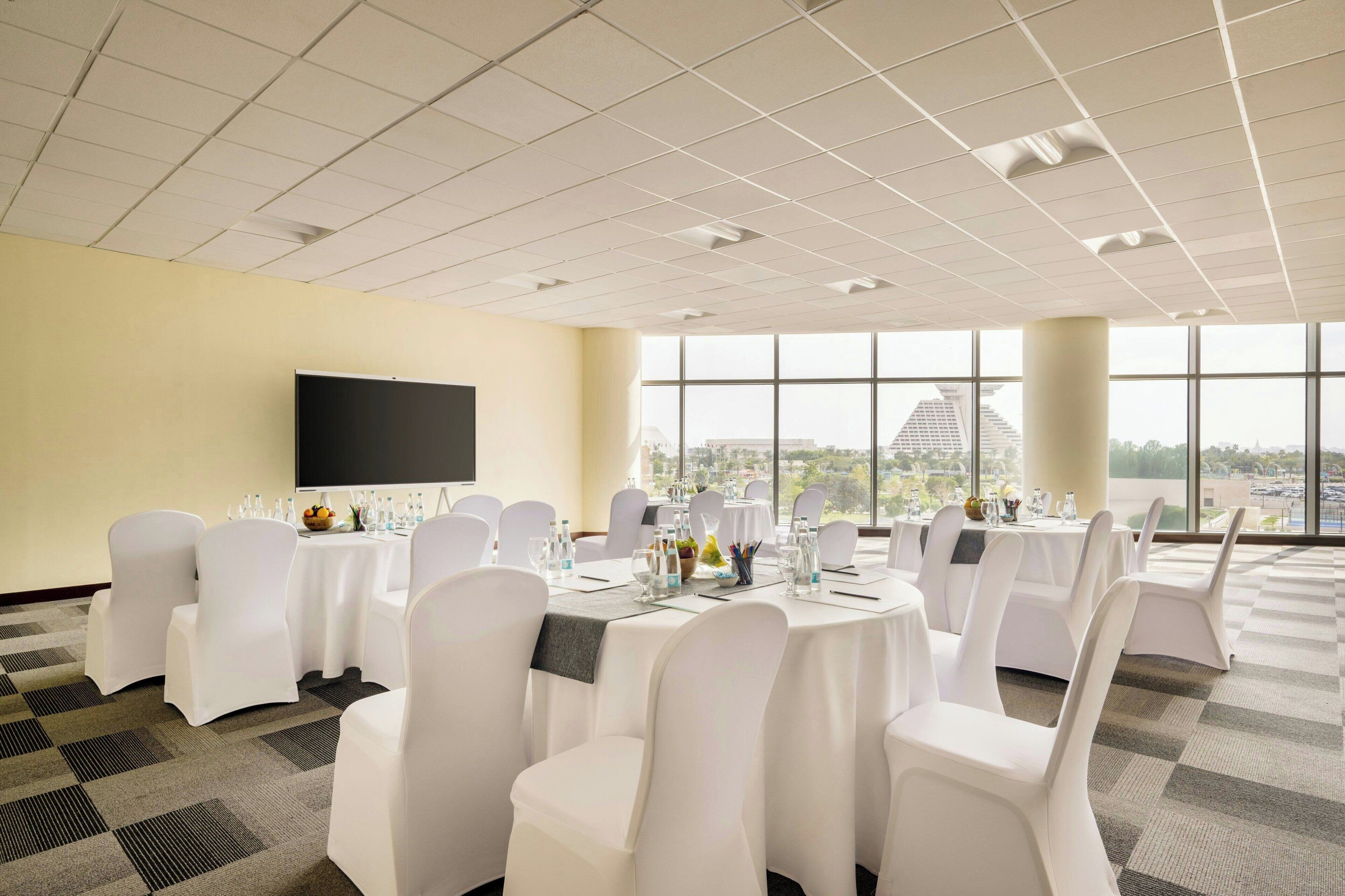 A conference room with round tables, white chairs, and a large screen on the wall.