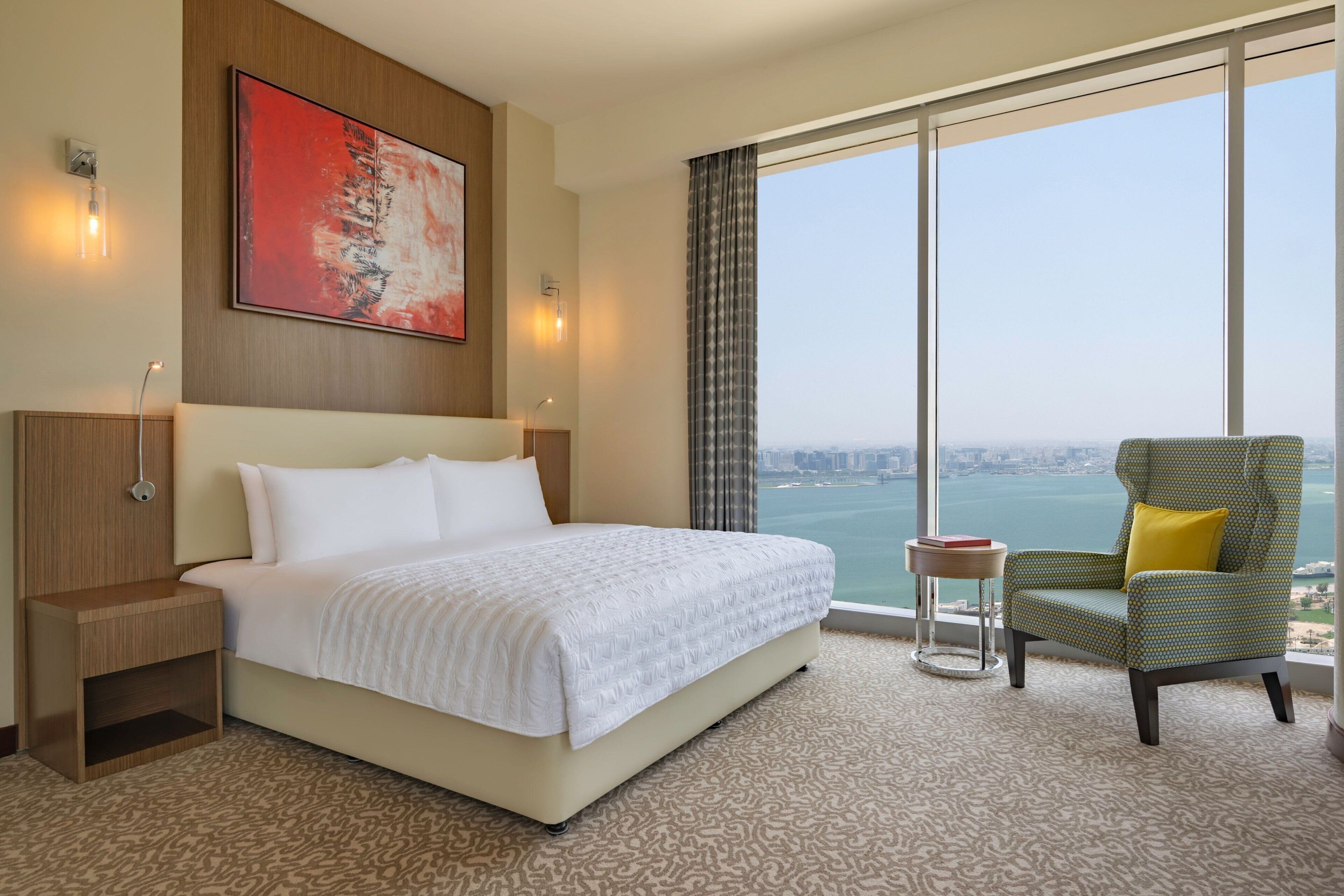 A hotel room with a large bed, a view of the water, a chair, and a painting on the wall.