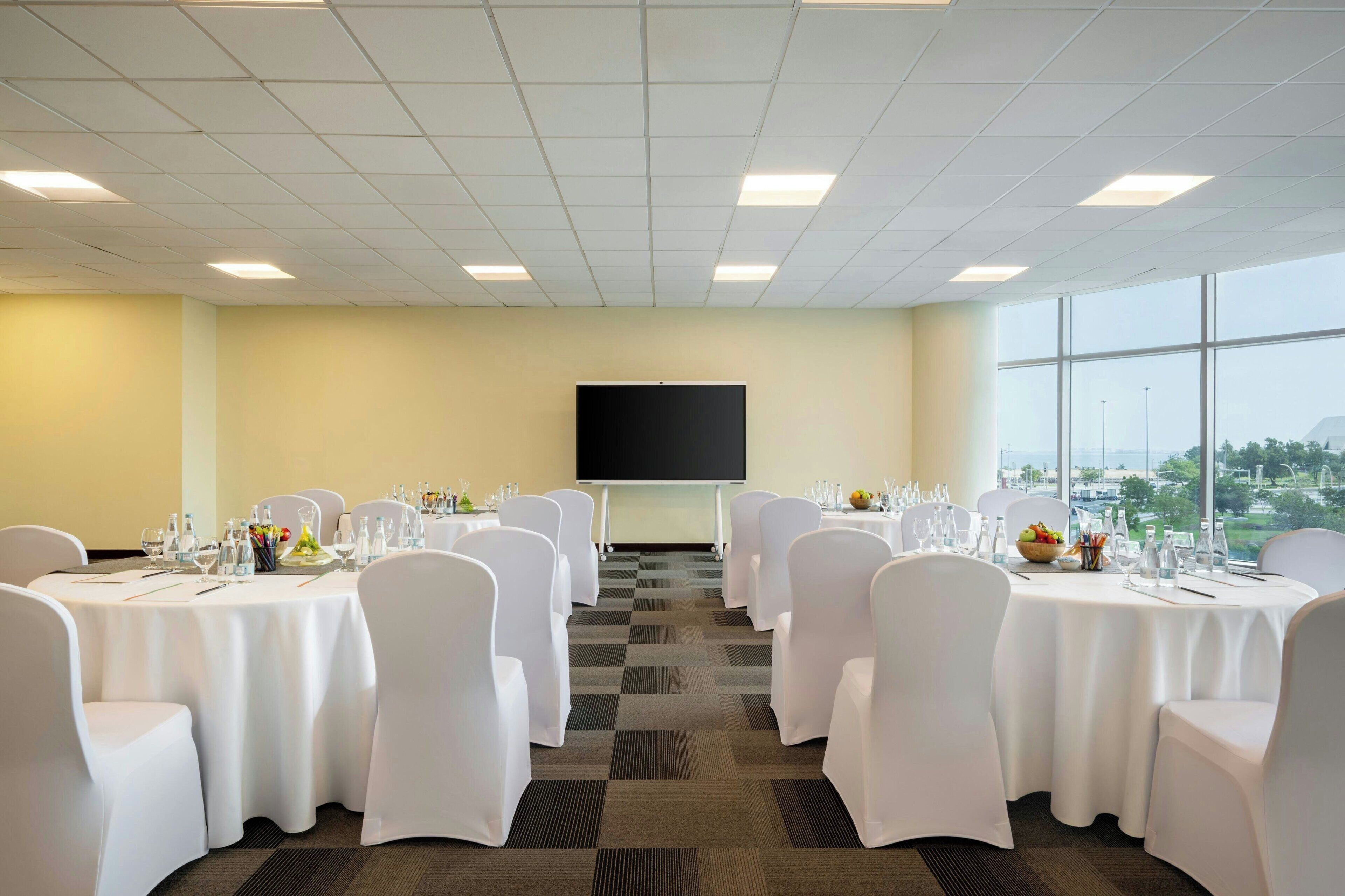 A conference room with white chairs and tables, a projector screen, and large windows with a view.