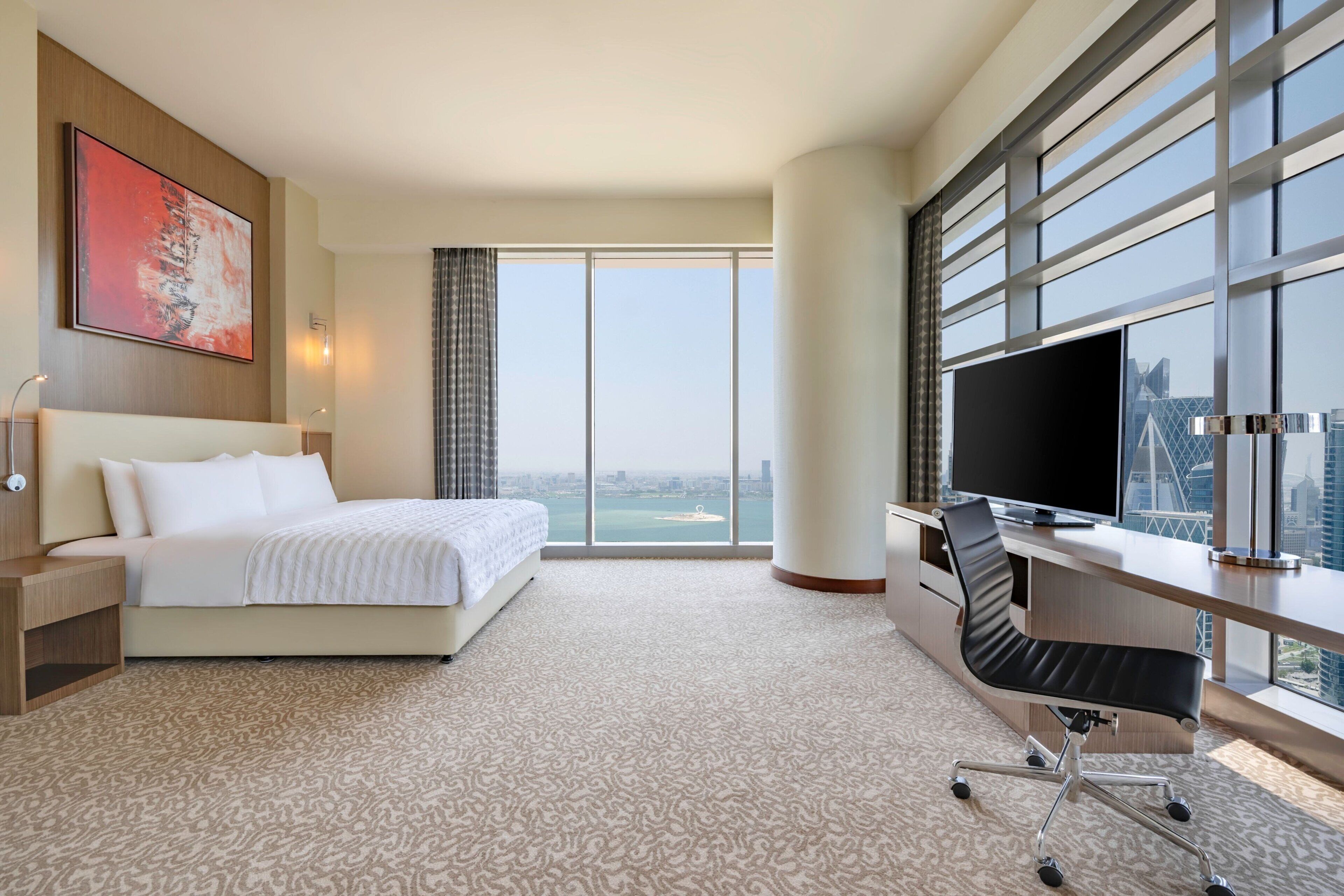 Een hotelkamer met een bed, een bureau met een computer, een televisie en grote ramen met uitzicht op de stad.