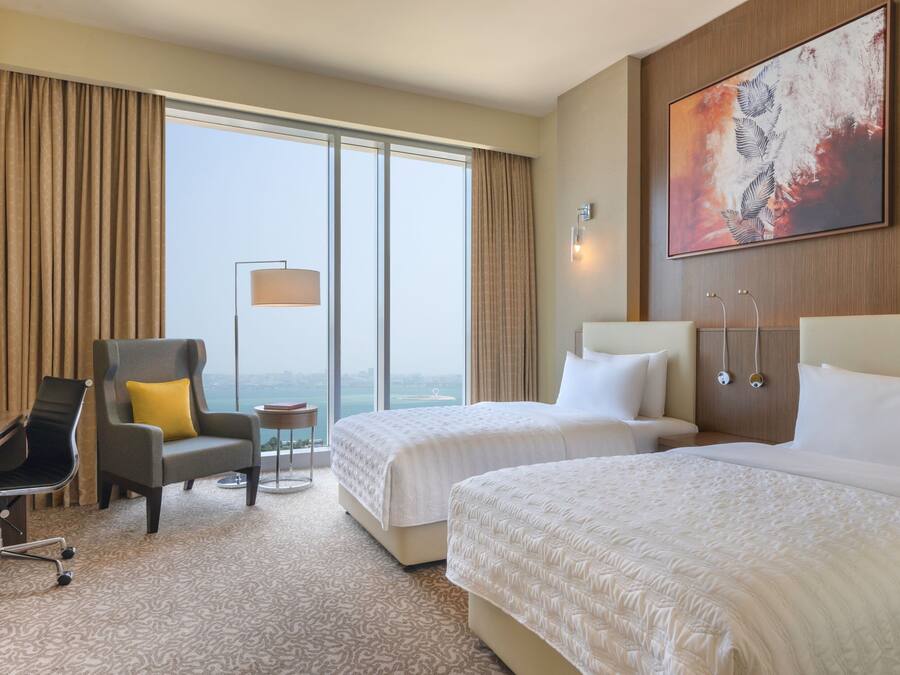 A hotel room with two beds, a desk, a chair, a small table, and a large window with curtains.