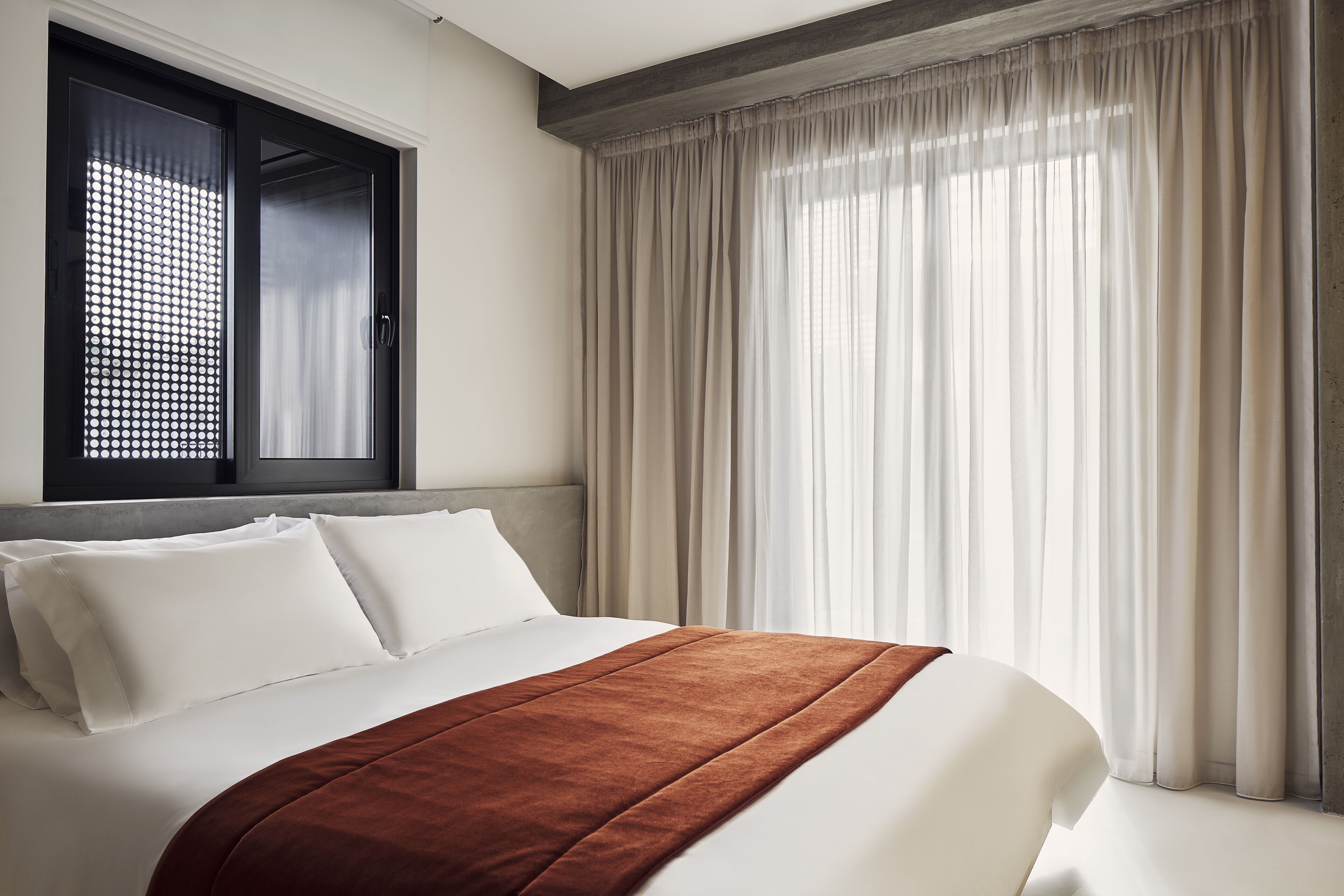 A hotel room with a bed, white bedding, a brown throw blanket, a window with sheer curtains, and a view of the outdoors.