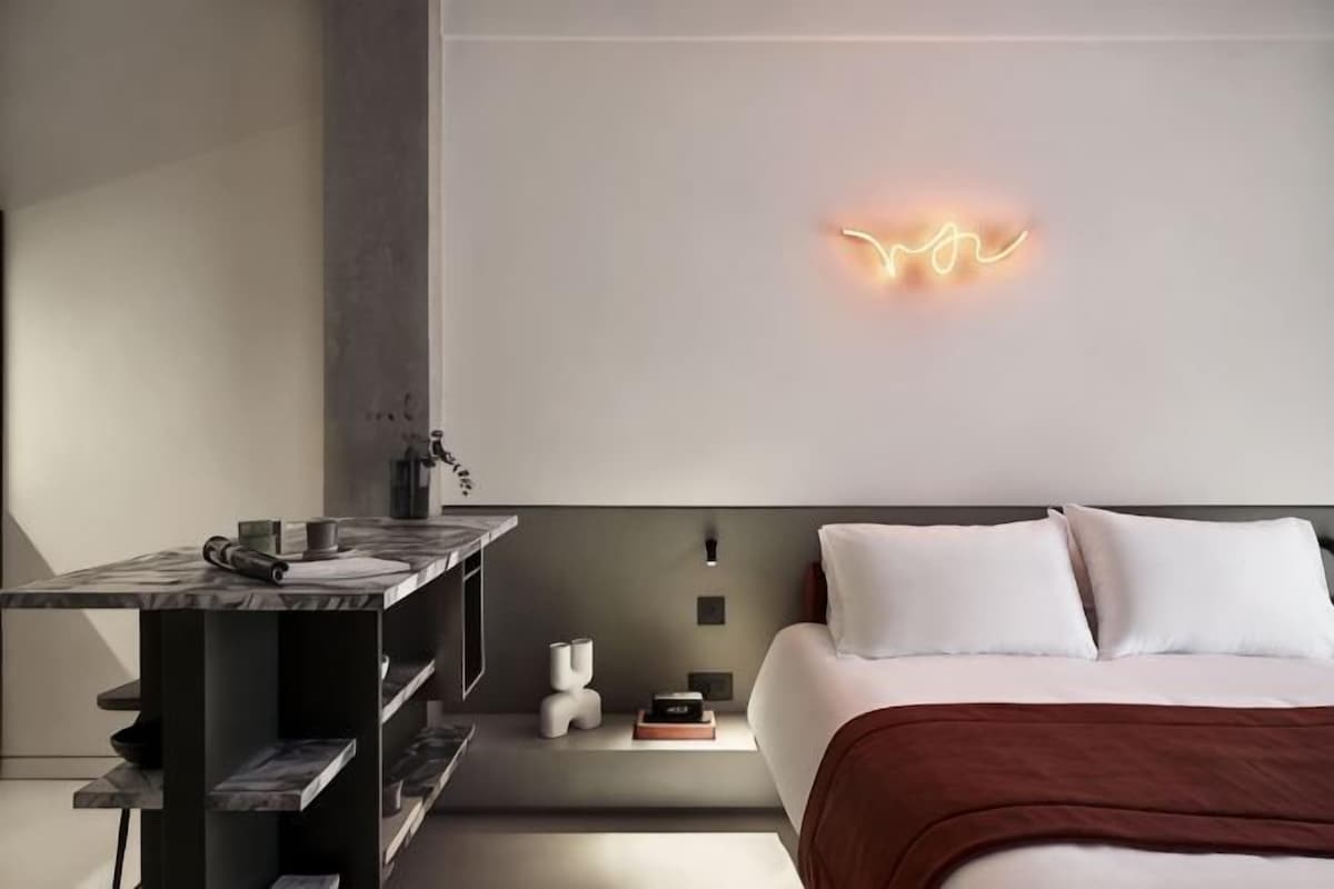 A modern hotel room with a bed, bedside table, and a wall-mounted light fixture.
