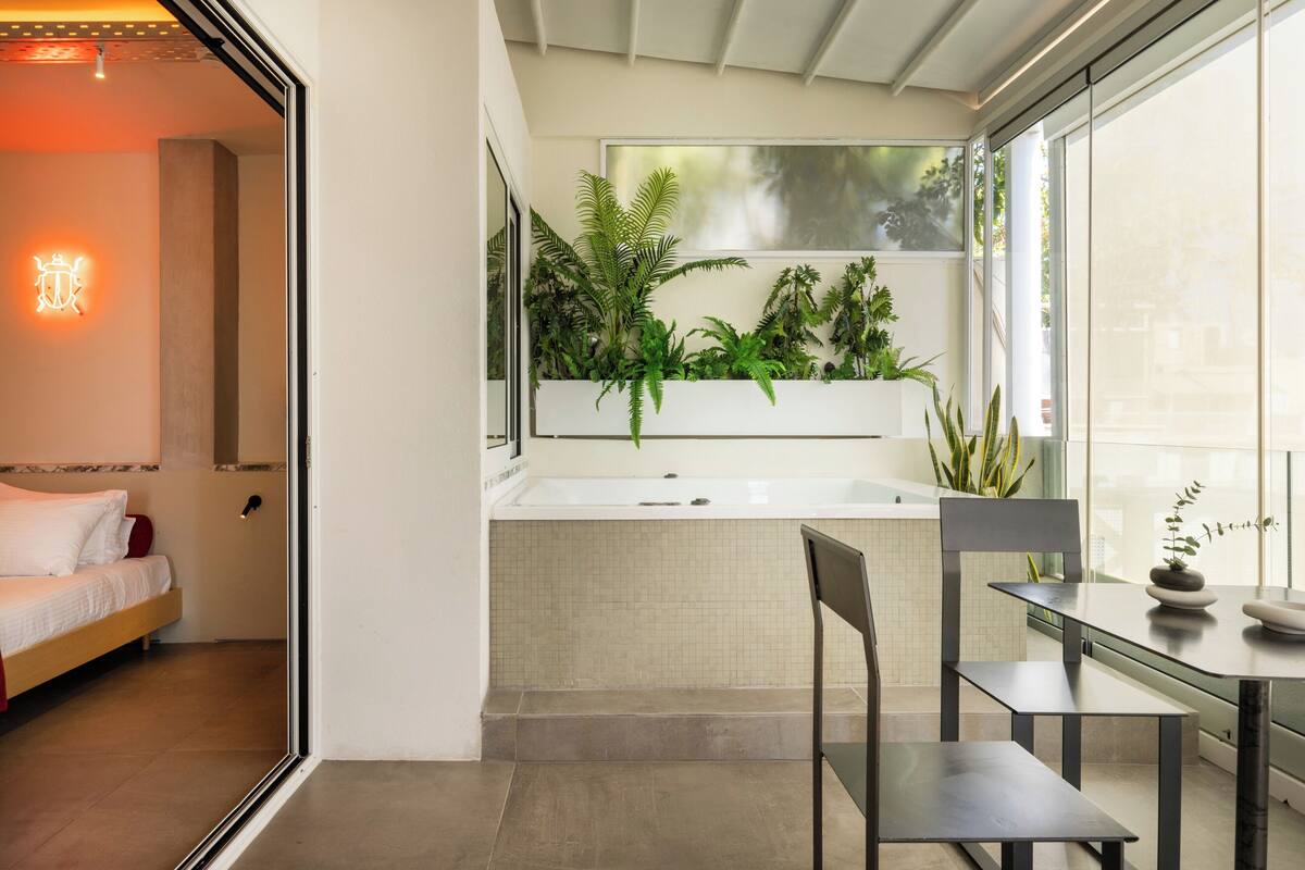 A modern bathroom with a large window, a bathtub, and a sink. There is a bed and a chair in the adjacent room.