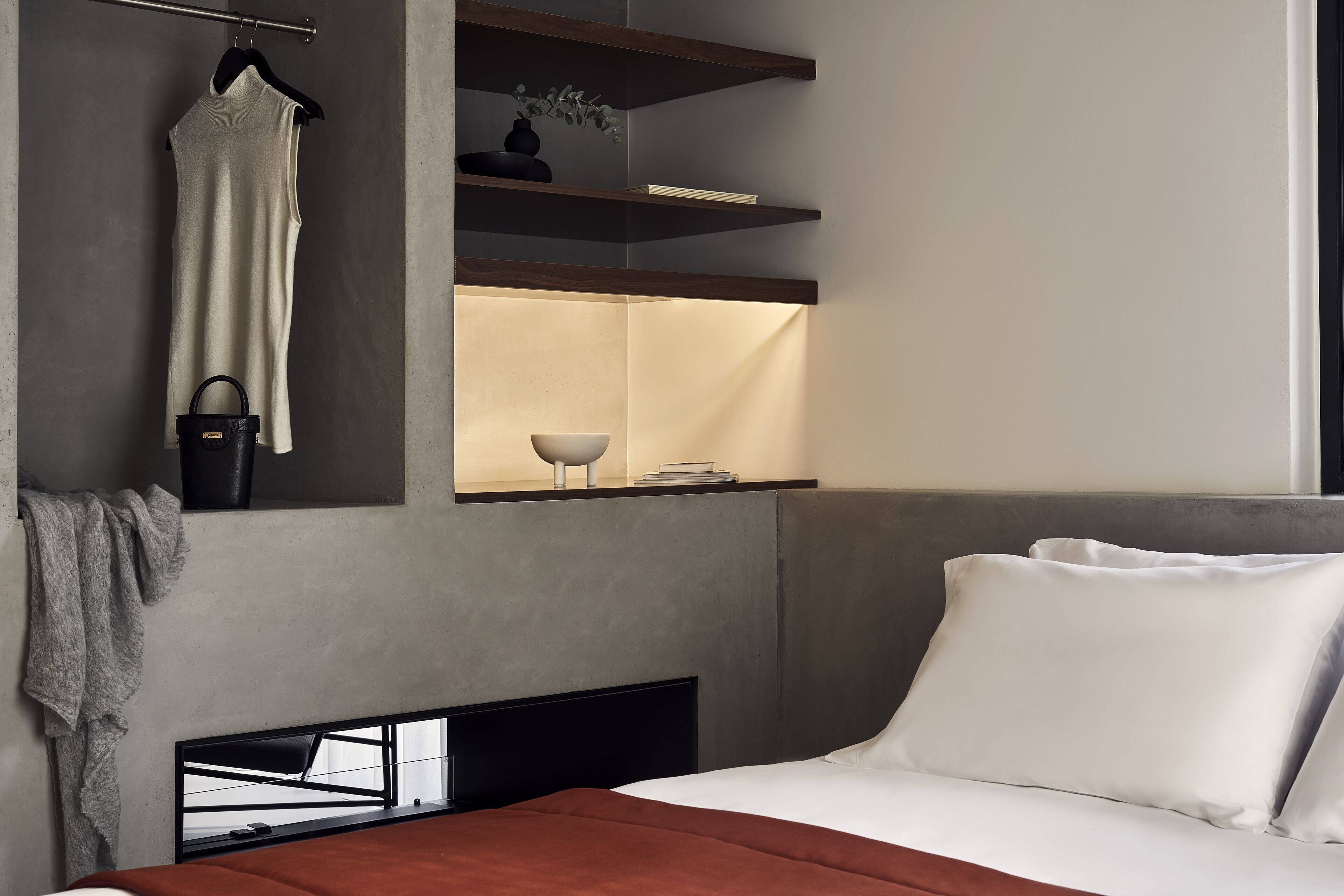 A modern hotel room with a bed, a hanging garment, a shelf with a vase, and a bedside table with a lamp.