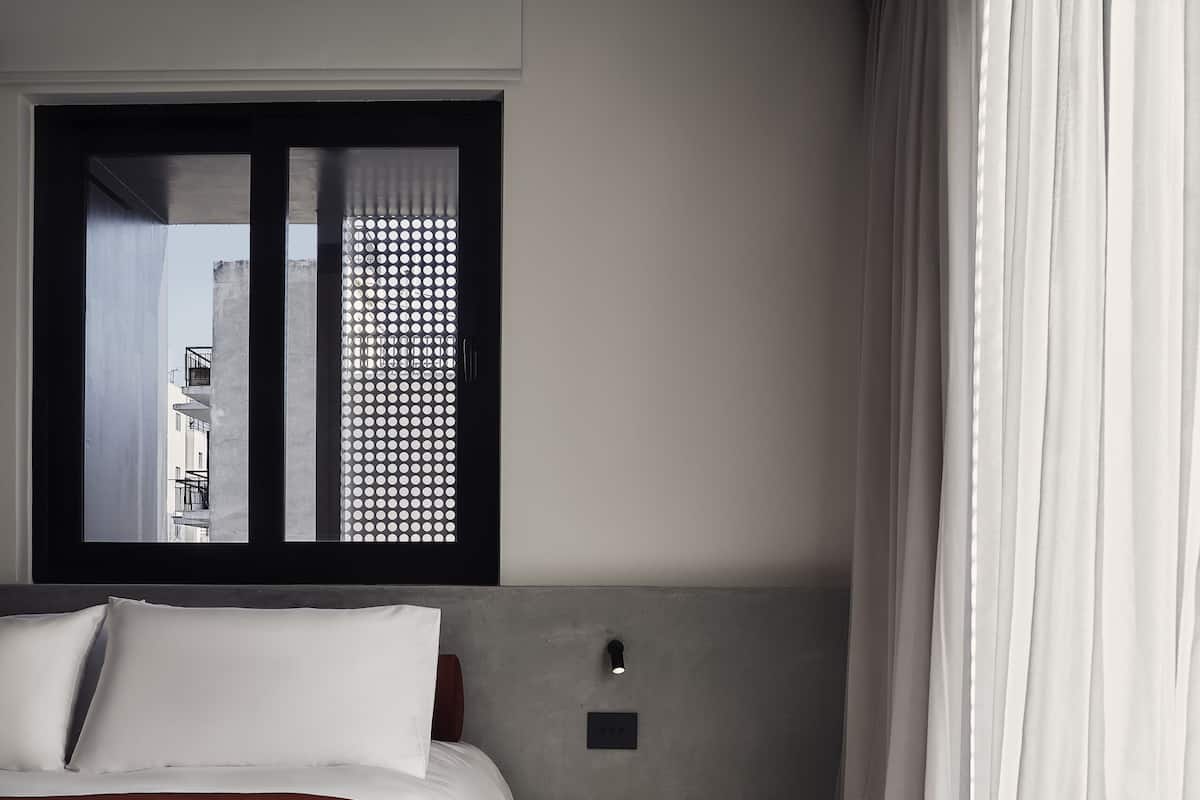 A modern hotel room with a large bed, a small bedside table, a window with a view of buildings, and a curtain.