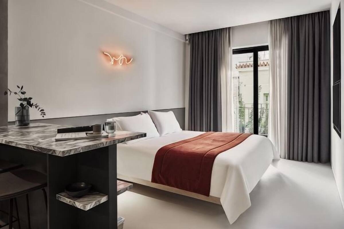 A modern hotel room with a bed, a desk, and a neon sign on the wall.