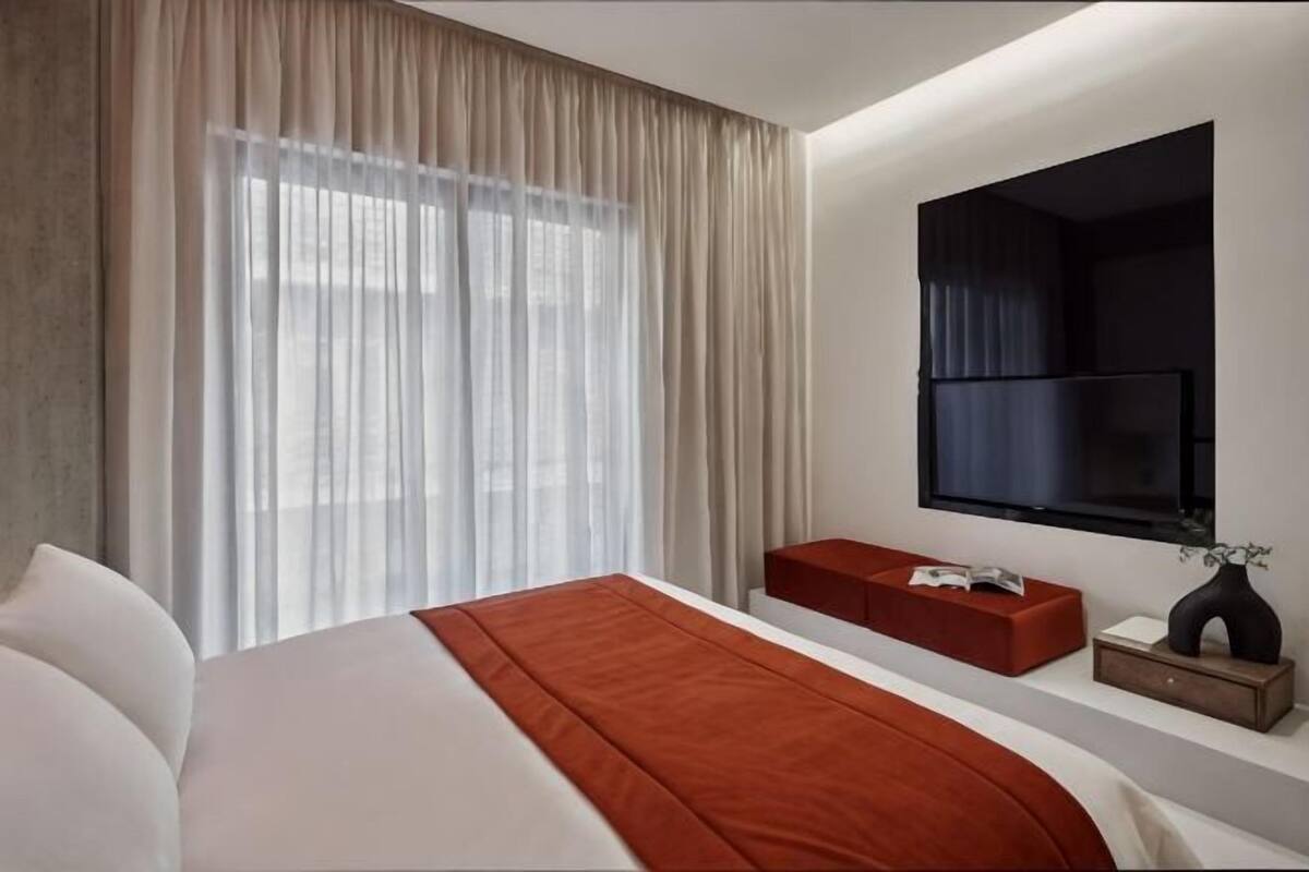 A hotel room with a bed, a TV mounted on the wall, a window with curtains, and a small table with a vase.