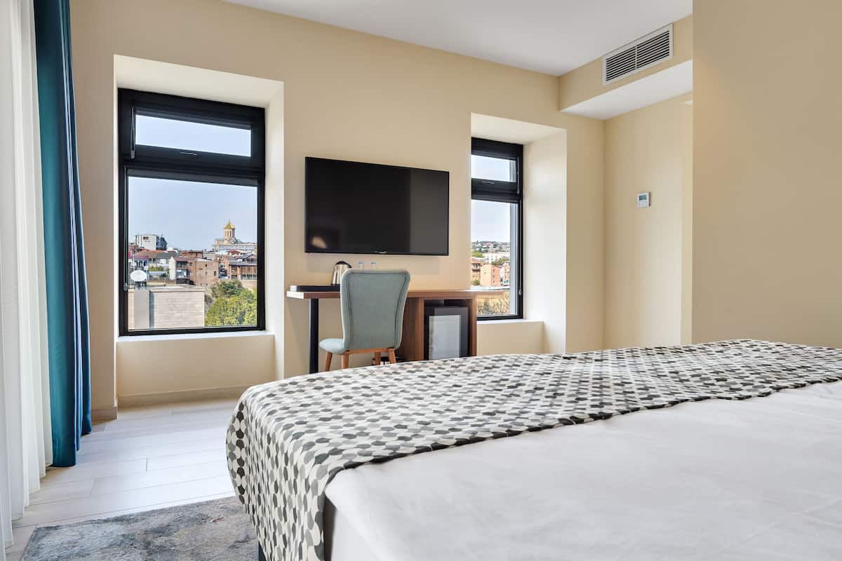 A hotel room with a large bed, a desk with a chair, a TV, and large windows offering a city view.