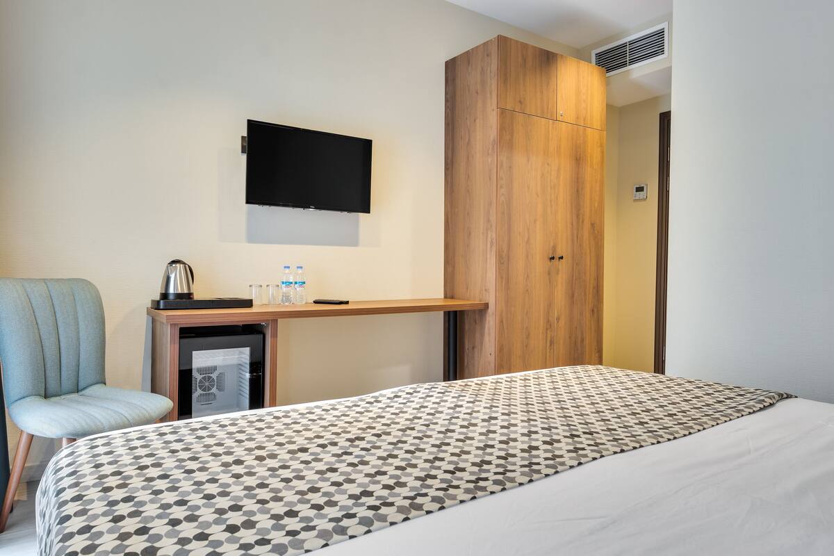 A hotel room with a bed, a desk with a flat-screen TV, a wooden wardrobe, and a chair.