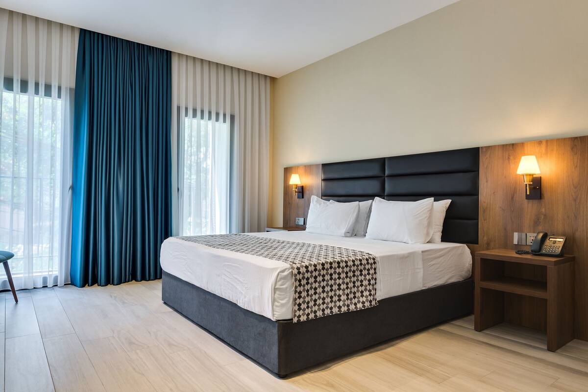 A modern hotel room with a large bed, two bedside tables, a wooden headboard, and a view of the outdoors through large windows.
