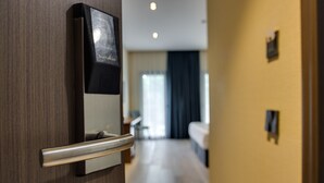 Entree van een hotelkamer met een digitaal slot en een deurklink, die uitkomt op een gang met een bed en een raam met gordijnen.