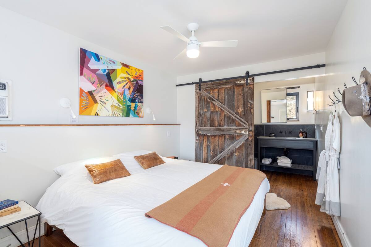 A bedroom with a large bed, a ceiling fan, a colorful abstract painting, a wooden door, a vanity with a mirror, and a robe hanging on the wall.