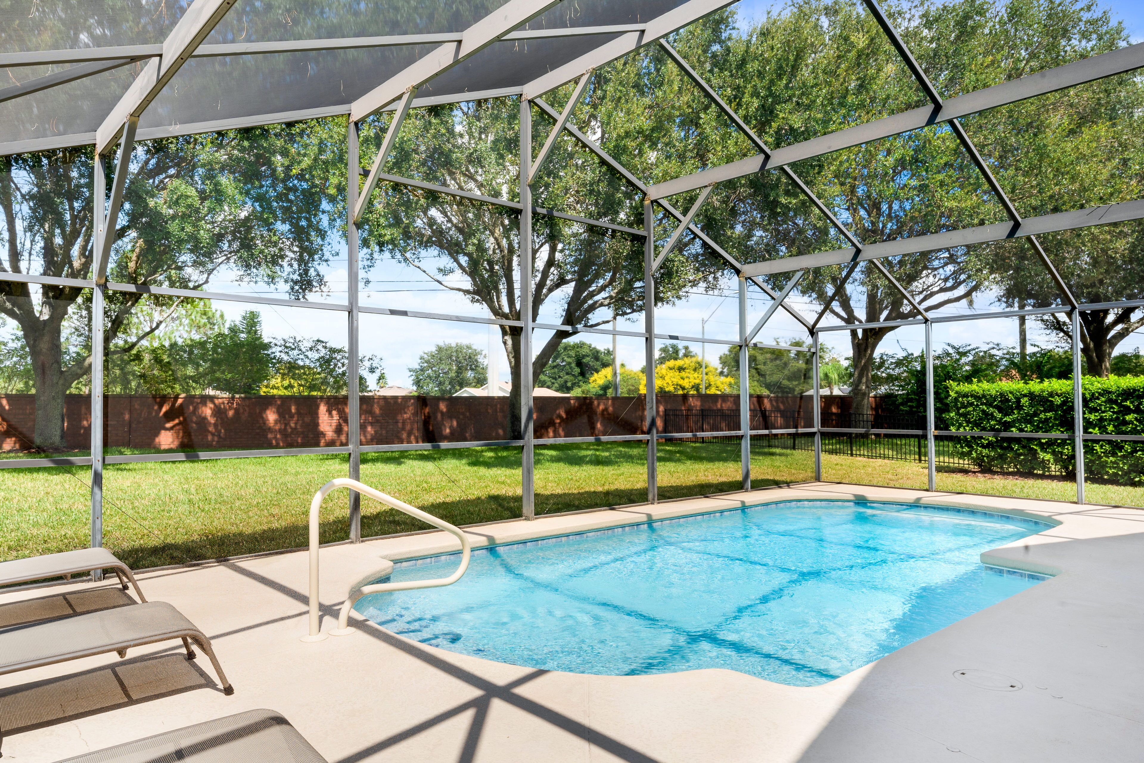 A covered outdoor swimming pool with a metal frame and glass panels, surrounded by a grassy area and trees.
