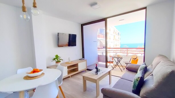 A modern living room with a sofa, a dining table with chairs, a TV mounted on the wall, and a balcony with a table and chairs. - Delight Holiday Home (Playa del Inglés)
