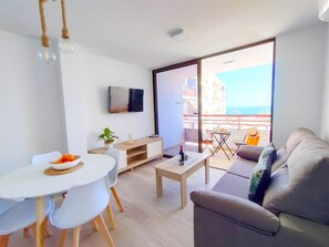Una sala moderna con sofá, una mesa de comedor con sillas, un televisor montado en la pared y un balcón con mesa y sillas.