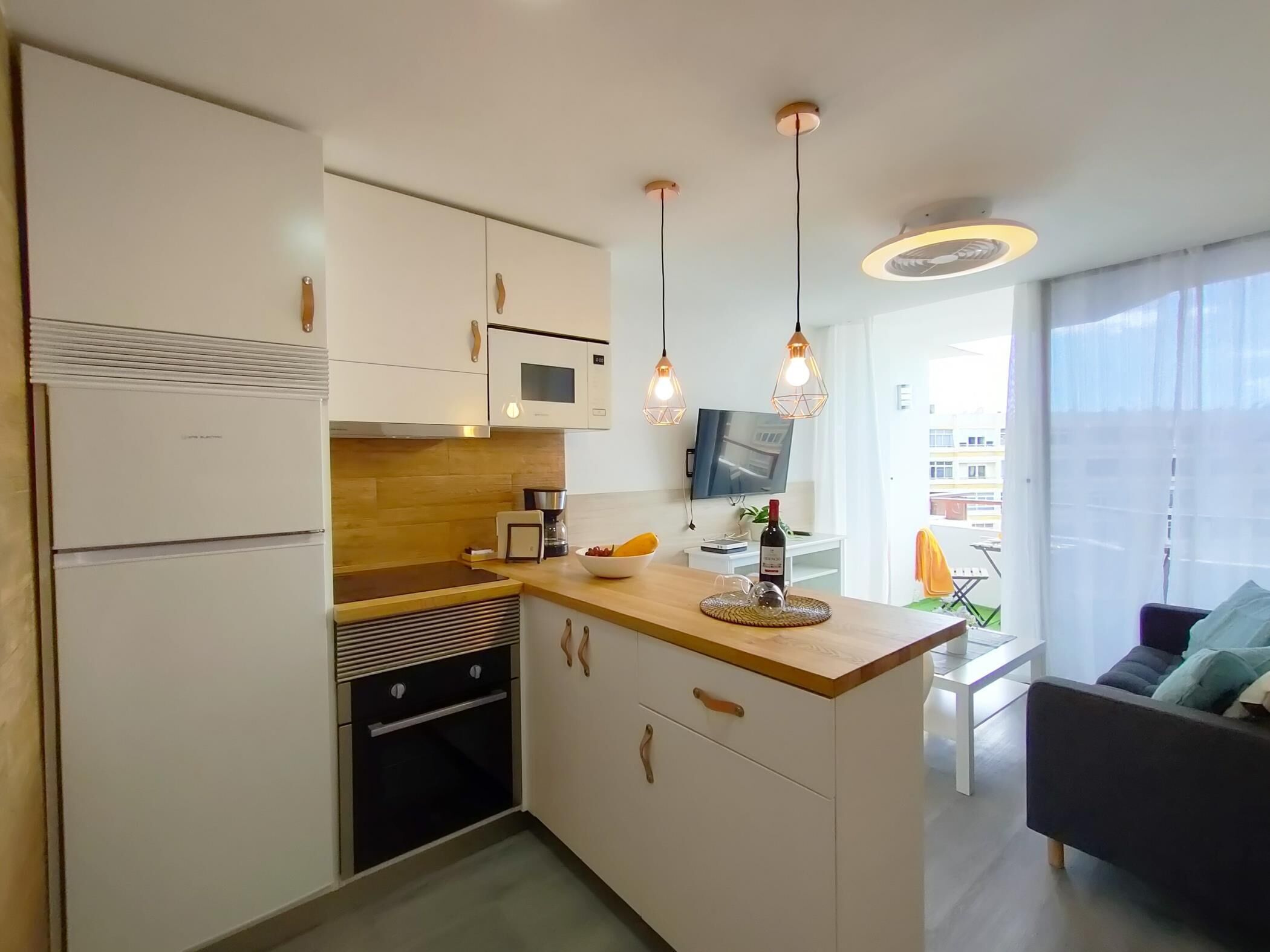 A modern kitchen with white cabinets, a microwave, a coffee maker, and a wine bottle on the counter. There is a wooden island with a bowl and a glass pitcher. A TV is mounted on the wall, and a sofa is visible in the background.