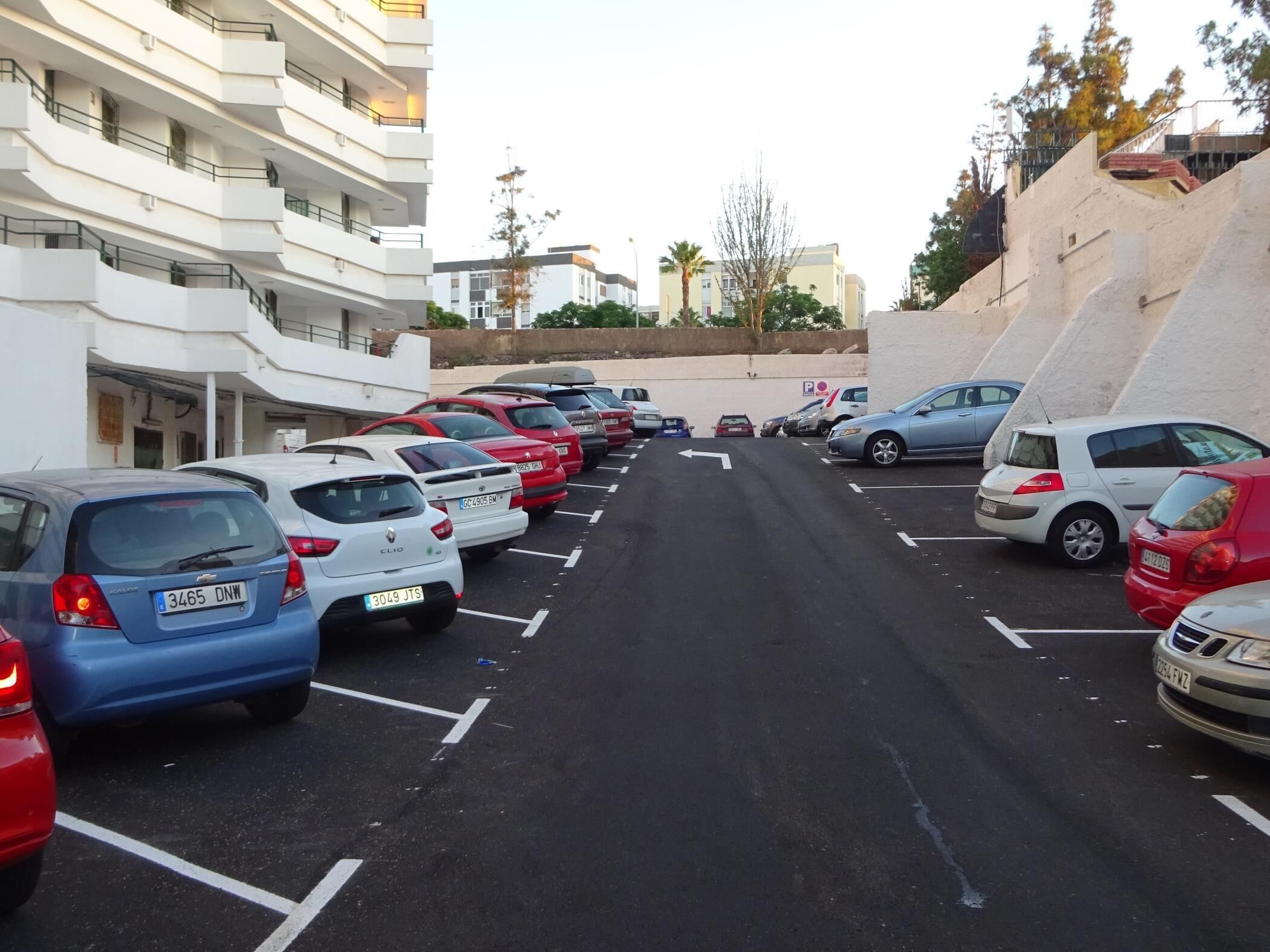 Un estacionamiento con varios coches aparcados en filas, un edificio con balcones a la izquierda y una pared inclinada a la derecha.