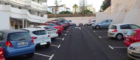 Un estacionamiento con varios coches aparcados en filas, un edificio con balcones a la izquierda y una pared inclinada a la derecha.