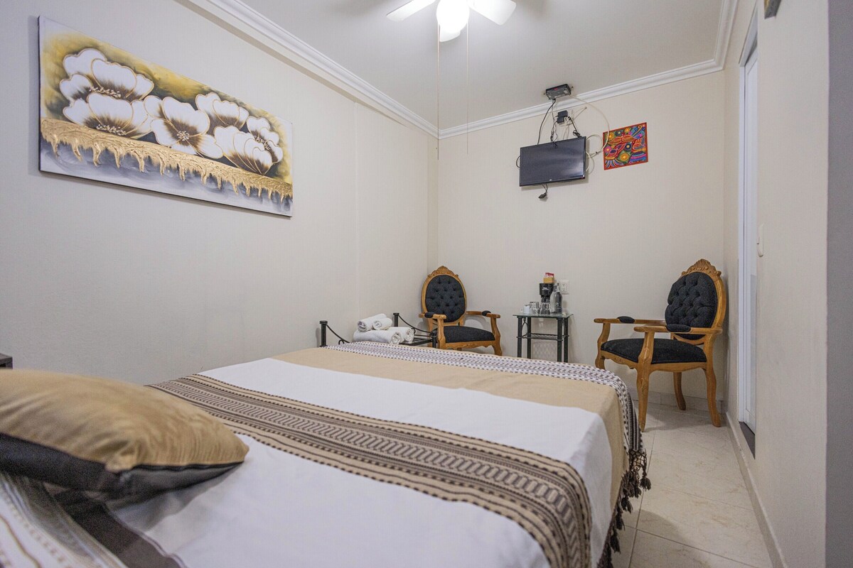 Una habitación de hotel con una cama, dos sillas, una mesita, una televisión y obras de arte en las paredes.