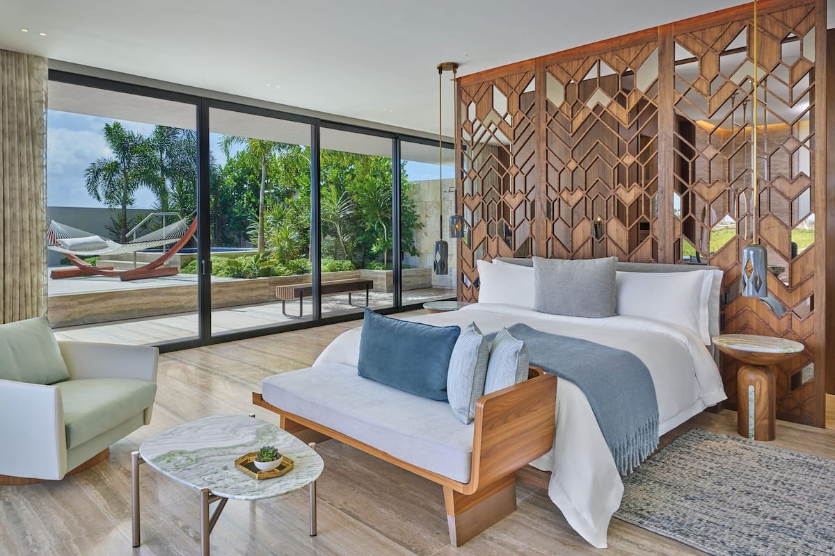 A modern bedroom with a large bed, a sofa, and a small table. There is a wooden partition with a geometric pattern and a view of greenery outside.