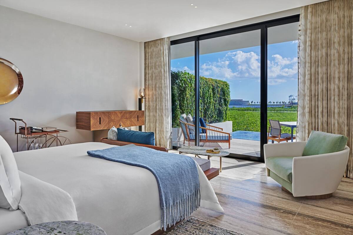 A modern hotel room with a large bed, a seating area with a table, and a view of a green landscape through large windows.