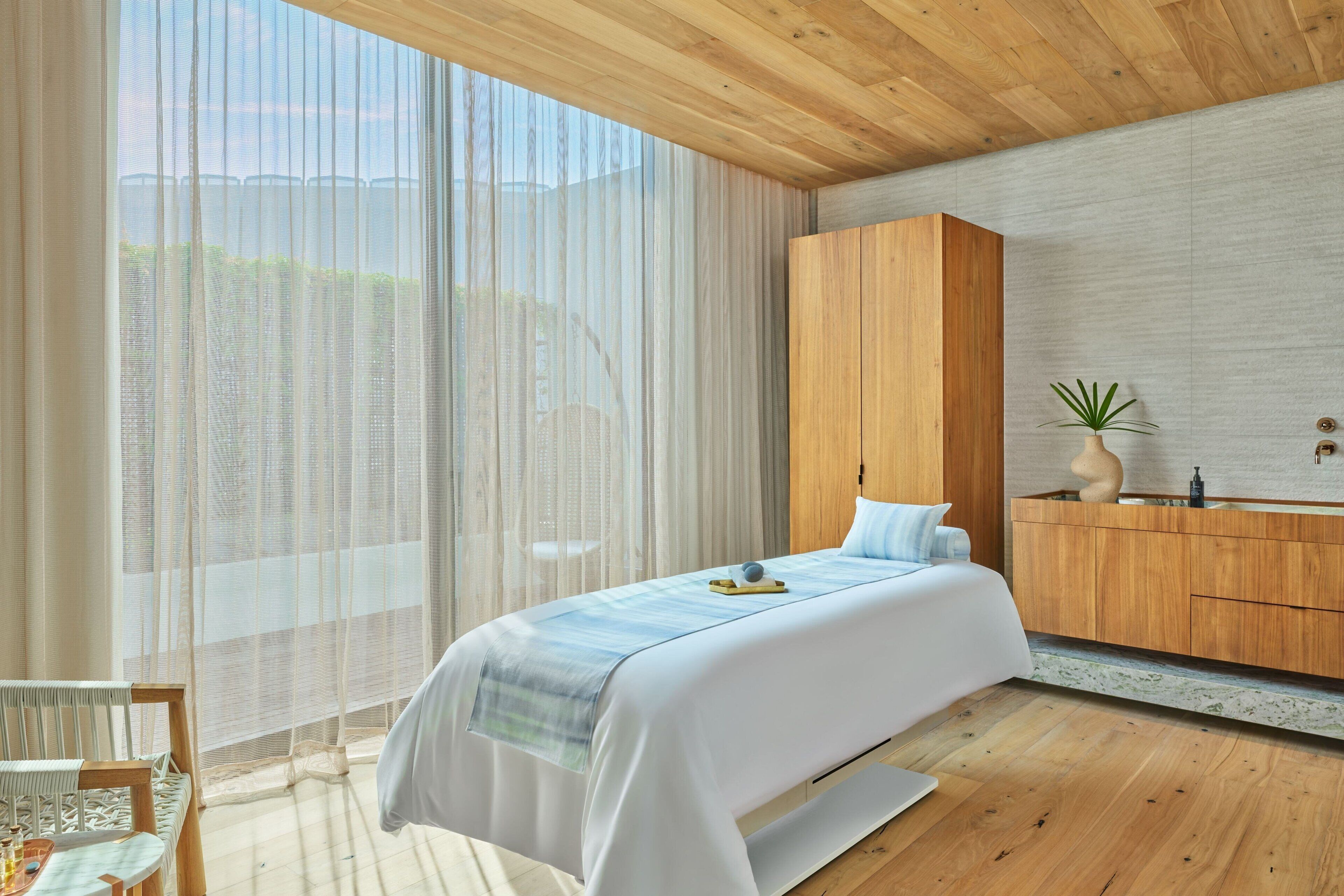 Une chambre moderne avec un plafond en bois, une grande fenêtre ornée de rideaux légers, un lit recouvert d’une couvre-lit à rayures bleues et blanches, une armoire en bois et une petite plante posée sur la commode.