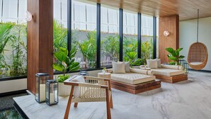 A modern living room with a sofa set, a rattan chair, and a hanging chair. Large windows provide a view of greenery outside.