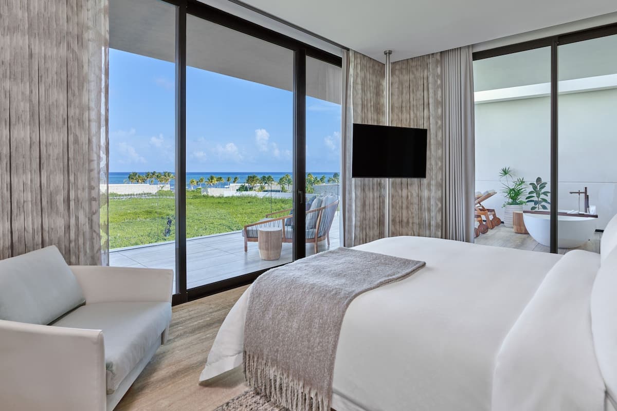 A modern hotel room with a large bed, a TV, and a balcony with a view of the beach and ocean.