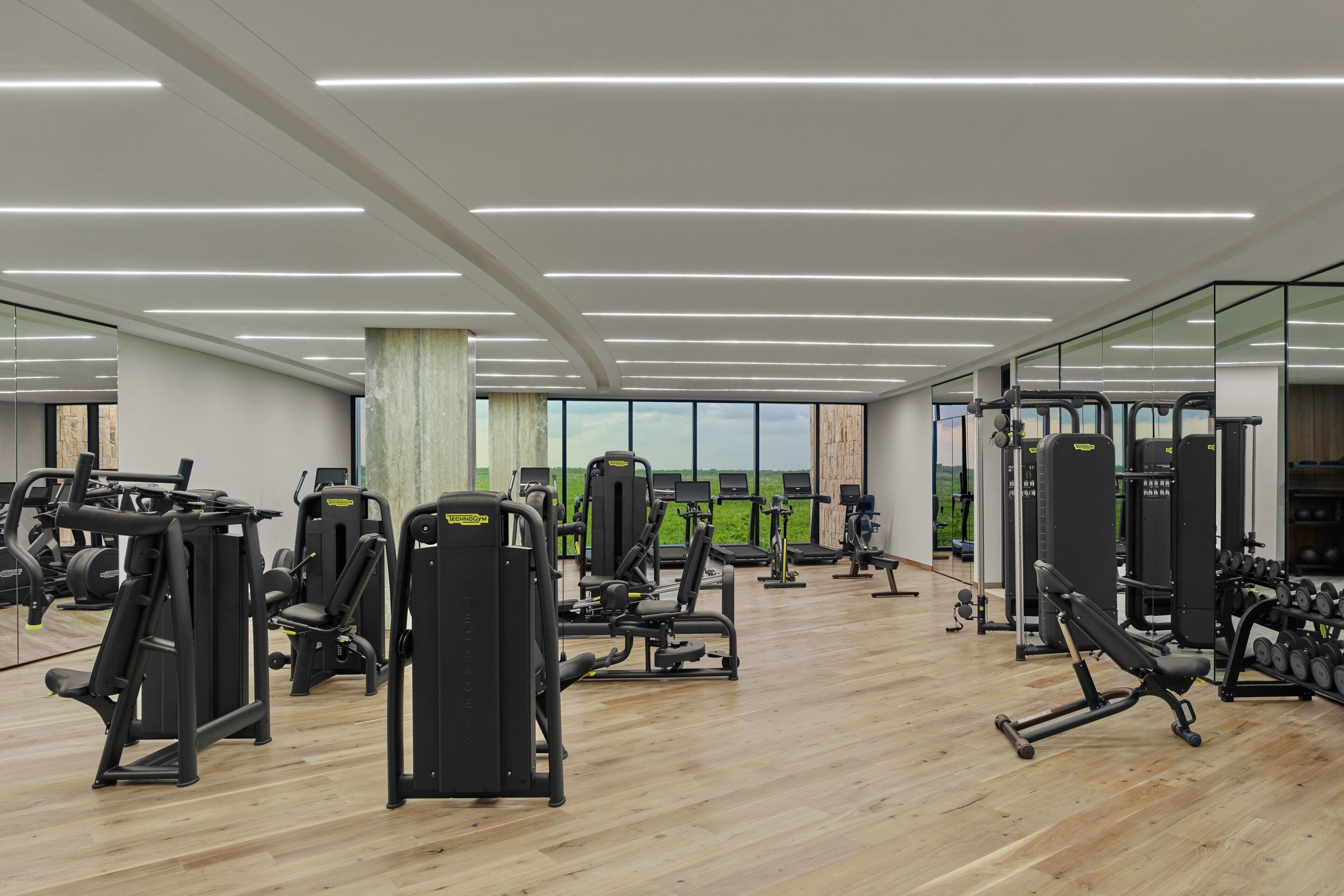 Uma academia com diversos equipamentos de exercício, janelas amplas e piso de madeira.