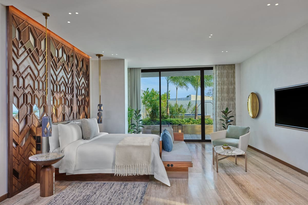 A modern bedroom with a large bed, a TV, a seating area, and a view of greenery outside.