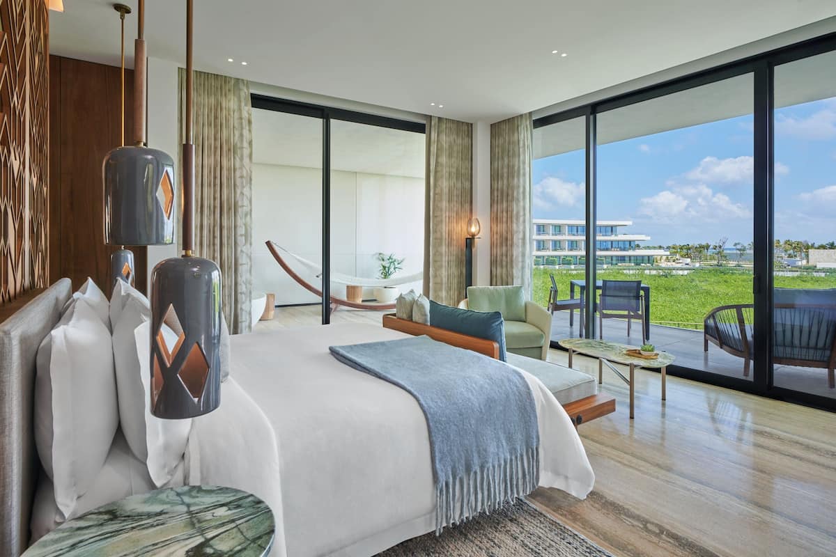 A modern hotel room with a large bed, a seating area with a table, and a view of a building and greenery outside.
