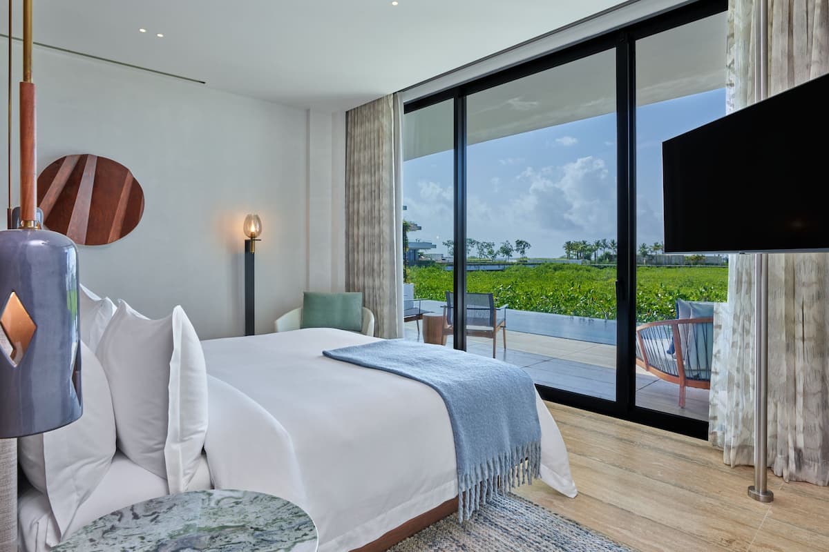 A modern hotel room with a large bed, a flat-screen TV, and a balcony with a view of the greenery and buildings.