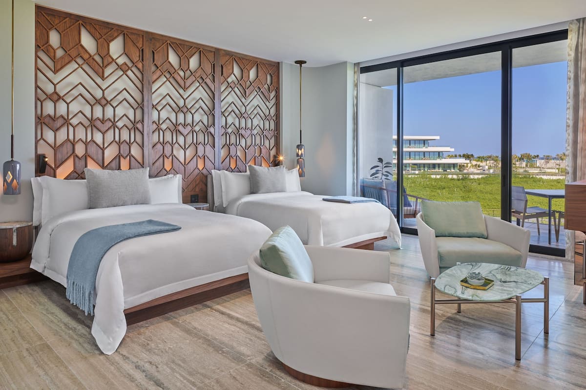 A modern hotel room with two beds, a seating area, and a large window offering a view of a building and greenery.
