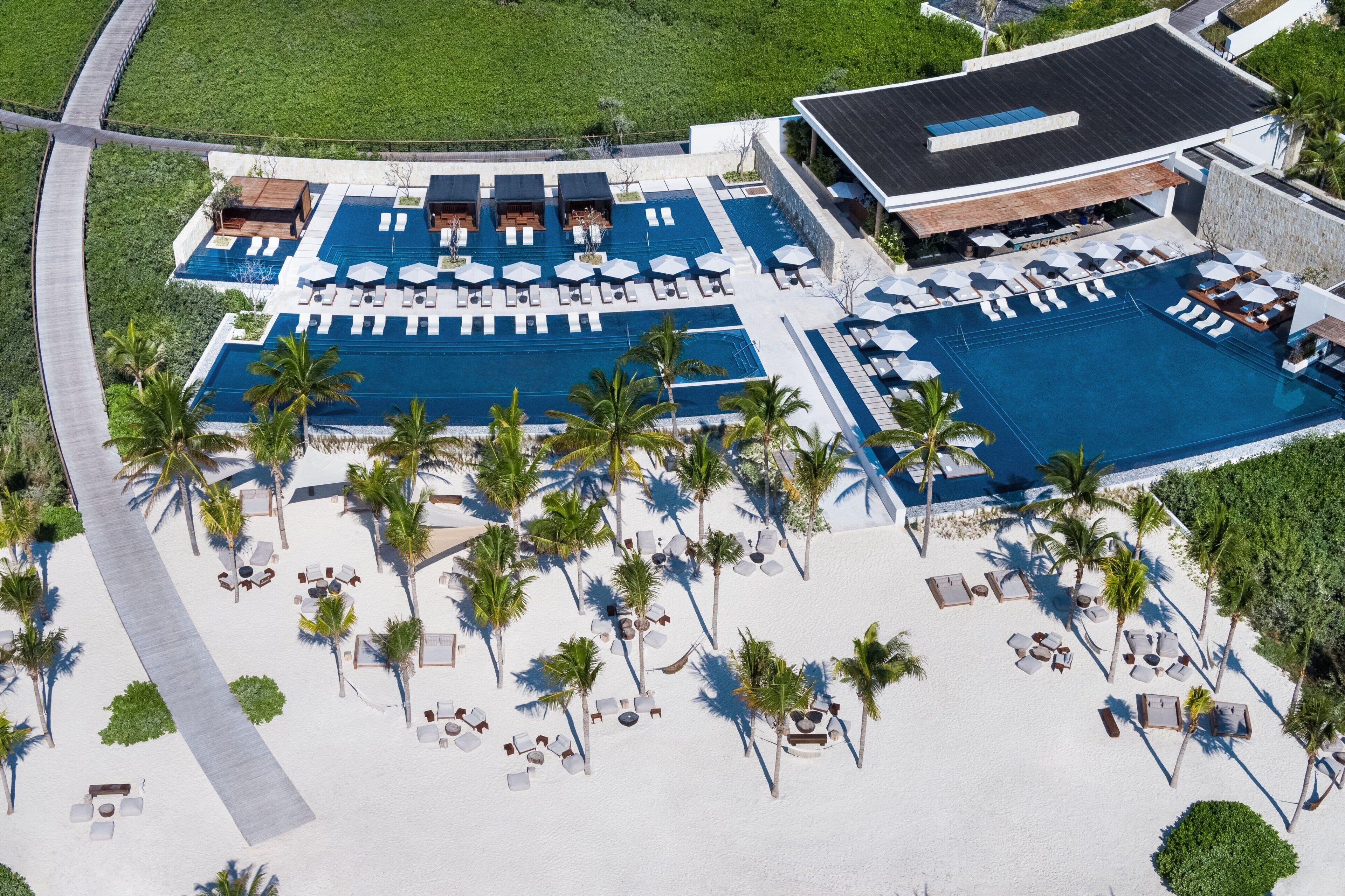 An aerial view of a resort with a large swimming pool, multiple lounge areas, and a beach area with sun loungers and palm trees.