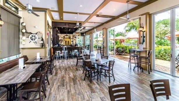 A restaurant with wooden flooring, tables, and chairs. It has a ceiling with wooden beams and a large window on the right side.