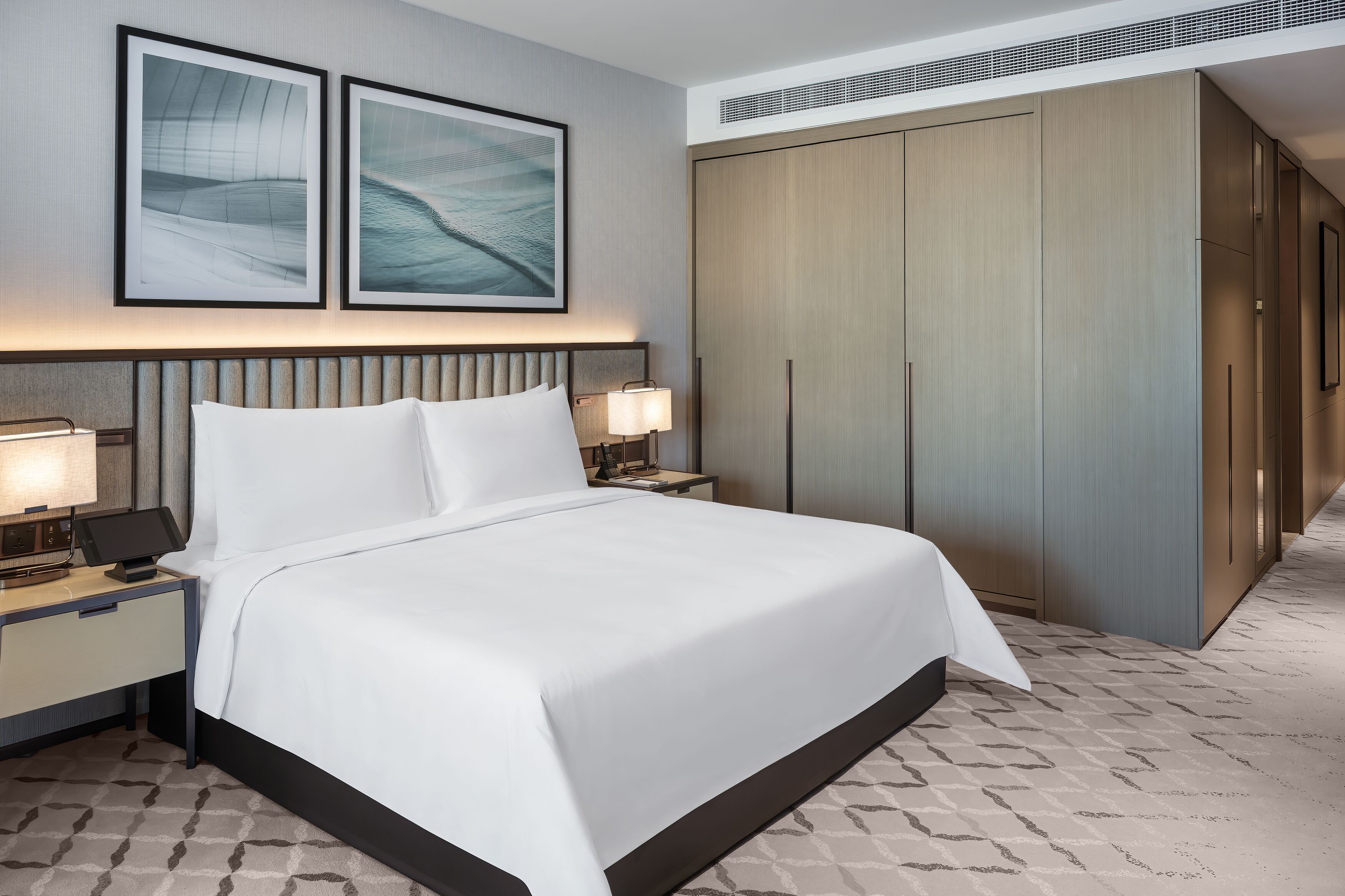 A hotel room with a large bed, two bedside tables with lamps, a wardrobe, and two framed artworks on the wall.