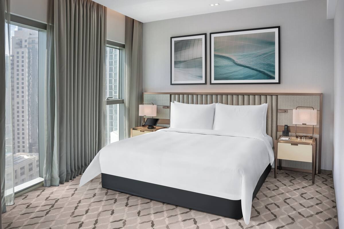 Habitación de hotel con una cama grande, dos mesitas de noche con lámparas, vistas de la ciudad a través de amplias ventanas y dos cuadros en la pared.