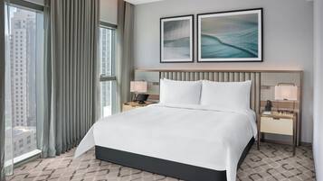 A hotel room with a large bed, two bedside tables with lamps, a view of the city through large windows, and two framed artworks on the wall.