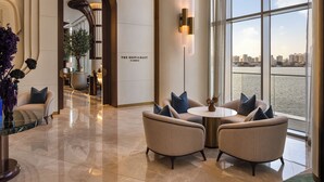 Un moderno vestíbulo de hotel con vista a un río y al horizonte de la ciudad, que incluye una zona de estar con sofás y una mesa, y un letrero que dice "The Restaurant at Address".