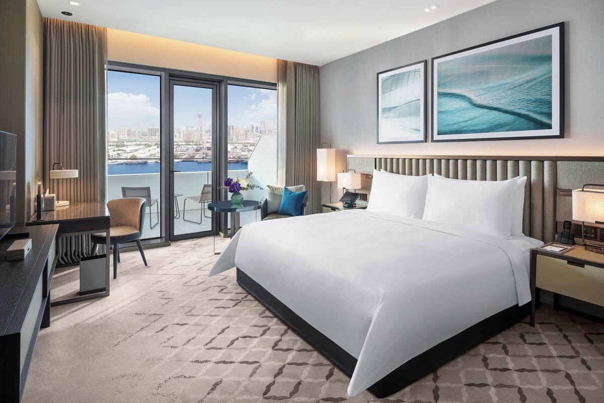 Habitación de hotel con una cama grande, un escritorio, una silla, una mesita con un florero y vistas a la ciudad a través de amplios ventanales.