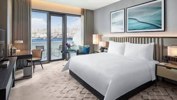 A hotel room with a large bed, a desk, a chair, a small table with a vase, and a view of the city through large windows.