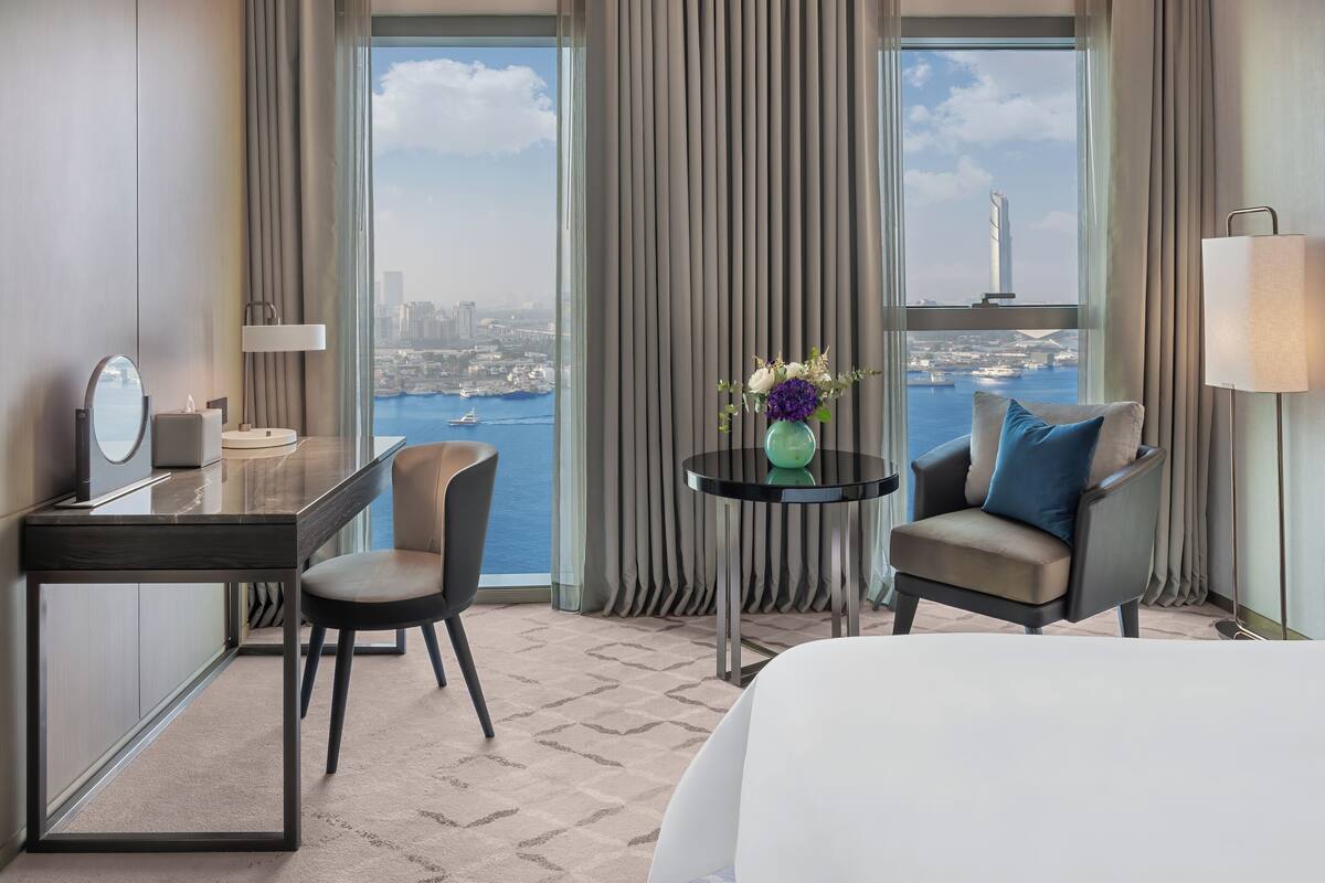Habitación de hotel con cama, escritorio, silla, una mesita con un florero y vistas a la ciudad y al agua.