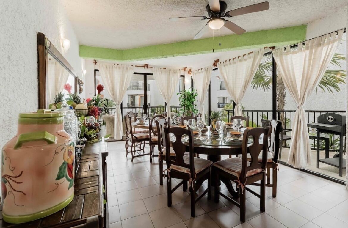 A dining area with a round table set for four, surrounded by wooden chairs. A ceiling fan is visible above, and the room has a balcony with curtains.