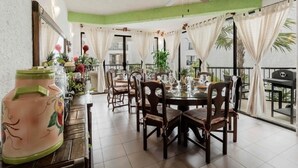 Un comedor con una mesa redonda dispuesta para cuatro personas, rodeada de sillas de madera. Se observa un ventilador de techo, y la habitación tiene un balcón con cortinas.