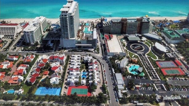 Vista aérea de un complejo turístico frente al mar, con varias construcciones, una piscina y una cancha de tenis.