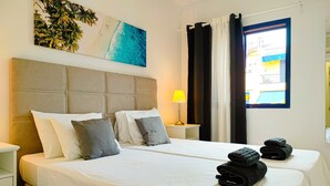 A hotel room with a large bed, two bedside tables, a nightstand, a lamp, a window with curtains, and a painting on the wall. - Lemon Terraced House (Playa del Inglés)