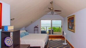 Una camera da letto con un letto grande, un ventilatore a soffitto, vista sul mare tramite una porta scorrevole in vetro e un tavolino con un oggetto decorativo.